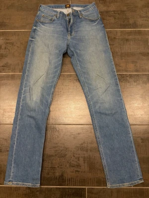 Blå raka jeans från Lee - Klassiska blå jeans från Lee med rak passform och fem fickor. Jeansen har normal midja, bälteshällor och dragkedjegylf. Tillverkade i slitstarkt denimtyg som ger en tidlös look. Perfekta för dig som gillar enkel och clean stil.30/34