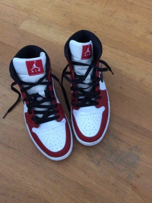 Nike Air Jordan 1 High röd/vit/svart - Säljer ett par Nike Air Jordan 1 High sneakers i klassisk röd, vit och svart färg. Skorna har svart swoosh, svarta skosnören och perforerad tåbox. Ikonisk Jumpman-logga på plösen och Air Jordan-märke på sidan. Perfekta för dig som gillar streetwear och basketstil.