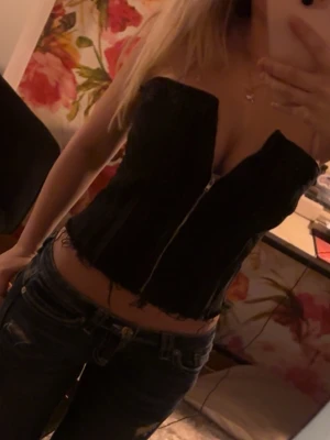 Svart korsett med dragkedja - Snygg svart korsett med dragkedja framtill och rå kant nertill. Toppen är axelbandslös och har en tight passform som framhäver midjan. Perfekt för att skapa en edgy look till jeans eller kjol.