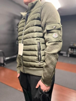 Olivgrön dunjacka från Moncler - Snygg olivgrön dunjacka från Moncler med quiltad front, ribbade muddar och ärmficka med logotyp. Jackan har hög krage, dragkedja framtill och två sidofickor med dragkedja. Perfekt för kalla vinterdagar och riktigt trendig look.