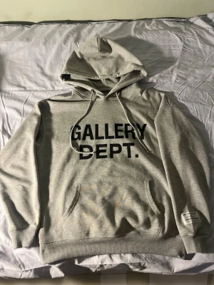 Grå hoodie från Gallery Dept. - Snygg grå hoodie från Gallery Dept. med svart tryck på bröstet. Klassisk huva med dragsnören och stor magficka framtill. Mjuk bomullsblandning och ribbade muddar vid ärmslut och nederkant. Perfekt för en avslappnad look. (Jag är osäker på äkthet eftersom jag köpt varan här på plick men jag tycker den ser äkta ut)