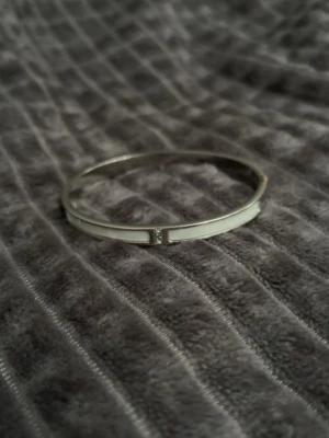 Silverfärgat stelt armband med vit detalj - Snyggt stelt armband i silverfärg med en vit, emaljliknande detalj längs utsidan. Armbandet har en minimalistisk och stilren design med en liten glittrande sten framtill. Perfekt för dig som gillar enkel men elegant stil.