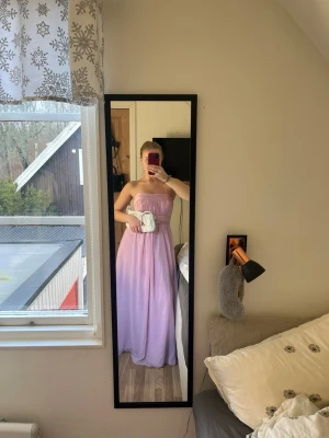 Ljusrosa balklänning  - Säljer en elegant ljuslila långklänning med axelbandslös design och markerad midja. Klänningen har ett flytande fall och är perfekt för speciella tillfällen exempelvis bal. 💕💕