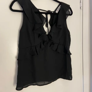 Svart volangtop med knyt i ryggen - Snygg svart ärmlös topp med volangdetaljer framtill och knyt i ryggen. Toppen har en lätt och luftig känsla, perfekt för att lyfta vilken outfit som helst. Passar dig som gillar feminina detaljer och stilren design.