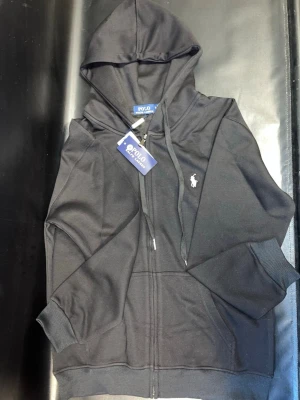 Svart hoodie från Polo Ralph Lauren - Svart hoodie från Polo Ralph Lauren med dragkedja framtill och klassisk huva. Märkets ikoniska logga i vitt är broderad på bröstet. Tillverkad i mjukt material och har ribbade muddar vid ärmar och nederkant. Perfekt för en avslappnad och stilren look.