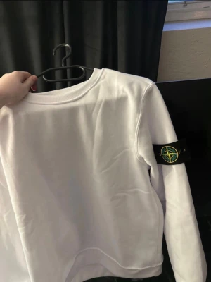 Vit sweatshirt från Stone Island - Säljer en stilren vit sweatshirt från Stone Island med klassisk logotyp-patch på vänster ärm. Tröjan har rund halsringning och ribbade muddar vid ärmslut och nederkant. Perfekt för en clean och modern look.