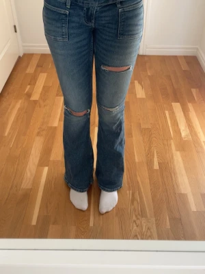 Blå bootcut jeans med slitningar - Säljer ett par blå bootcut jeans med slitningar över knäna. Jeansen har klassisk femficksmodell, normal midja och är tillverkade i ett mjukt denimtyg. Perfekta för en avslappnad och trendig look.