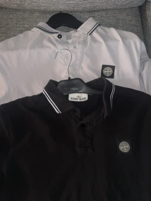 Pikétröjor från Stone Island - Två klassiska pikétröjor från Stone Island, en svart och en vit. Båda har krage med kontrastfärgade ränder och den ikoniska Stone Island-loggan på bröstet. Tillverkade i mjuk bomull för en bekväm och stilren look.