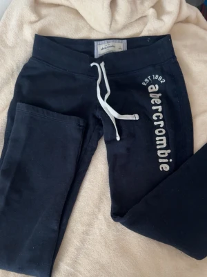 Mörkblå mjukisbyxor Abercrombie känns som XS - Snygga mörkblå mjukisbyxor från Abercrombie med vit dragsko i midjan och tryckt logga längs benet. Byxorna har en avslappnad passform och är tillverkade i mjukt bomullsmaterial. Perfekta för chill dagar eller när du vill vara bekväm.