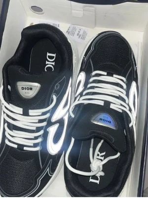 Dior b30 - Snygga svarta Dior sneakers med vita reflekterande detaljer och vita skosnören. Skorna har en sportig design med mesh och syntetmaterial, samt tydlig Dior-logga på plösen och innersulan. Perfekta för dig som vill sticka ut med exklusiva sneakers. Pris kan diskuteras. Svarar snabbt!