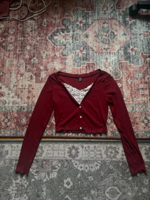 Vinröd croppad cardigan med spets - Vinröd croppad cardigan med långa ärmar och vita knappar framtill. Tröjan har en snygg spetsdetalj i vitt vid urringningen och vågig kant nedtill. Knappt använd 