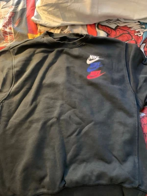 Svart stickad sweatshirt från Nike - Grön stickad sweatshirt från Nike med tre Nike-loggor i vitt, blått och rött på bröstet. Tröjan har rund halsringning och ribbade muddar vid ärmslut och nederkant. Perfekt för en avslappnad streetwear-look.