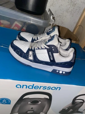 Louis Vuitton blåvita sneakers - Snygga sneakers från Louis Vuitton i blått och vitt med klassiska LV-loggor på sidan och hälen. Skorna har lågt skaft, vita snören och detaljer i mörkblå mocka och läder. Perfekta för dig som vill sticka ut med exklusiv design och streetwear-vibe.