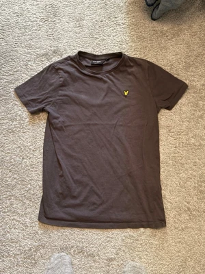 Brun t-shirt från Lyle & Scott - Säljer en grå t-shirt från Lyle & Scott med klassisk gul logga på bröstet. T-shirten har rund halsringning och korta ärmar. Tillverkad i mjuk bomull som känns skön mot huden. 