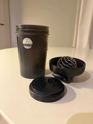 Svart shaker med metallvisp - Säljer en svart shaker i plast med tillhörande metallvisp och två lock. Perfekt för att blanda proteinshakes eller andra drycker. Märkt med 'BPA FREE GENUINE' på sidan. Enkel att ta med till gymmet eller skolan.