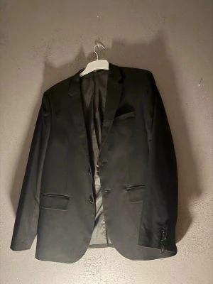 Svart klassisk kavaj med knappar - Stilren svart kavaj med två knappar framtill och klassiska fickor. Perfekt för dig som vill ha en tidlös och enkel look. Kavajen har lång ärm och slät finish, passar till många olika outfits.