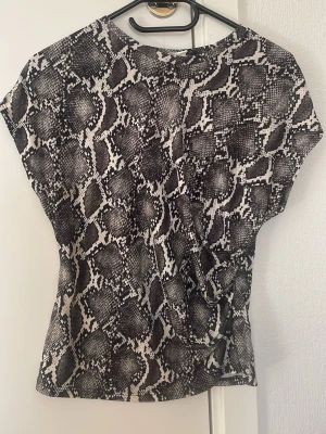 Grönvit  topp med ormmönster - Snygg topp med svartvitt ormmönster och korta ärmar. Toppen har en loose passform och är tillverkad i ett mjukt, stretchigt material som känns skönt mot huden. Perfekt för dig som vill sticka ut med ett djuriskt print.