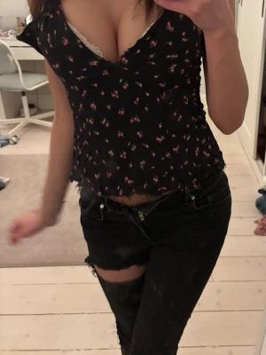 Svart blommig v-ringad blus - Söt svart blus med små blommor i rosa och rött. V-ringad med volangkant och lätt croppad passform. Perfekt för dig som gillar en feminin och trendig stil.