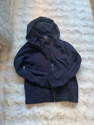 Mörkblå hoodie från Polo Ralph Lauren - Säljer en mörkblå hoodie från Polo Ralph Lauren med dragkedja och huva. Klassisk röd logga broderad på bröstet och mjukt material som känns skönt mot huden. Perfekt för dig som gillar stilrena och tidlösa plagg. 14/16 barn storlek 