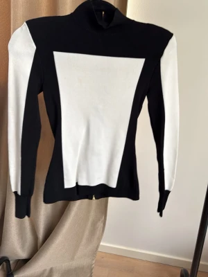 Svartvit tröja med grafiskt mönster Balmain x H&M - Säljer en svart och vit tröja med höghalsad krage och grafiskt mönster. Tröjan har långa ärmar och dragkedja i ryggen. Materialet är stretchigt och figurnära, perfekt för dig som gillar en modern och stilren look.