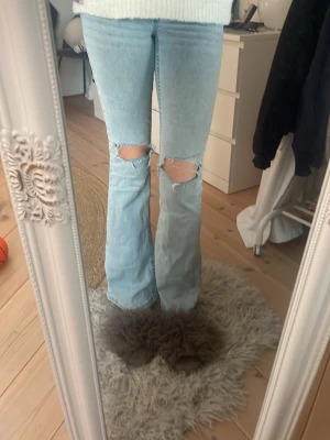 Ljusblå bootcut jeans med slitningar -  Säljer ett par ljusblå jeans med rosa detaljer på fickorna från Nelly. Det är bootcut och har en fin passform med ett midjemått rakt över på 32 cm och en inner benlängd på 82 cm. Orginlpris 699 kr.