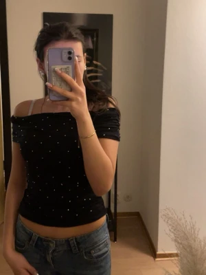 Svart glittrig offshoulder topp Zara - Svart offshoulder topp från Zara med glittriga prickar över hela tyget. Toppen är tight och croppad med korta ärmar och stretchigt material som sitter snyggt på kroppen. Perfekt för dig som gillar trendiga detaljer och en stilren look.