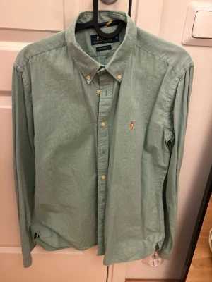 Grön skjorta från Polo Ralph Lauren - Snygg grön skjorta från Polo Ralph Lauren i custom fit. Skjortan har klassisk krage, knappar framtill och den ikoniska broderade loggan på bröstet. Tillverkad i mjuk bomull och har långa ärmar med knapp vid manschetten.
