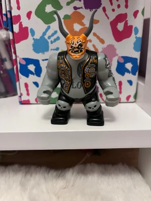 Lego-figur med horn och tatueringar - Unik Lego-figur med grå kropp, svarta byxor och orange mask med stora svarta horn. Figuren har detaljerade tatueringar på armarna och en väst med tryck. Perfekt för dig som gillar samlarfigurer eller vill bygga ut din Lego-värld.
