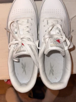 Vita Reebok Club C 85 sneakers - Klassiska vita Reebok Club C 85 sneakers i lågt snitt. Skorna har ovandel i slätt vitt läder, perforerade detaljer på tån och Reebok-logga med brittiska flaggan på sidan och plösen. Tjock vit sula och vita skosnören för en clean look. NÄSTINTILL HELT OANVÄNDA