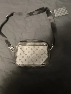 Louis Vuitton svart axelväska - Snygg svart axelväska från Louis Vuitton med klassiskt monogrammönster i grått och svart. Väskan har justerbar axelrem med präglad logga och en liten rutmönstrad nyckelring. Perfekt för dig som vill ha en stilren och trendig accessoar.