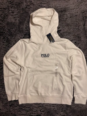 Vit hoodie från Polo Ralph Lauren - 18-20 Snygg vit hoodie från Polo Ralph Lauren med broderad logga på bröstet. Klassisk känguruficka framtill och ribbade muddar vid ärmslut och nederkant. Tillverkad i mjukt bomullsmaterial och har en stor huva för extra komfort.