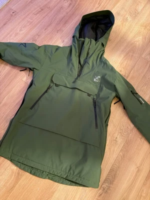 Grön skidjacka från RevolutionRace - Säljer en grön skidjacka från RevolutionRace med huva och flera dragkedjeförsedda fickor. Jackan har en stor bröstficka, justerbar huva och är tillverkad i slitstarkt syntetmaterial. 