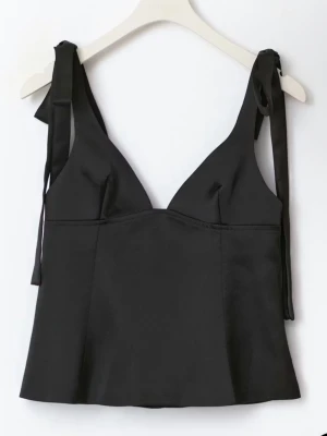 Svart satinlinnetopp med knytband - Stilren svart linnetopp i satin med smickrande v-ringning och breda axelband som knyts på axlarna. Toppen har en croppad passform och lätt glansigt material som ger en lyxig känsla. Perfekt att styla med jeans eller kjol för en trendig look.