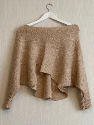 Beige stickad ponchotröja med glitter - Säljer en beige stickad ponchotröja med glittriga inslag. Tröjan har bred halsringning, asymmetrisk nederkant och långa ärmar med ribbade muddar. Perfekt för dig som gillar en avslappnad och trendig stil.