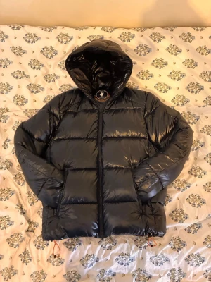 Svart Save The Duck pufferjacka - Säljer en svart Save The Duck pufferjacka med glansig finish och huva, nästan helt oanvänd Jackan har två sidofickor, dragkedja framtill och justerbar nederkant med dragsko. Märke med logga på vänster ärm. Perfekt för kalla vinterdagar.