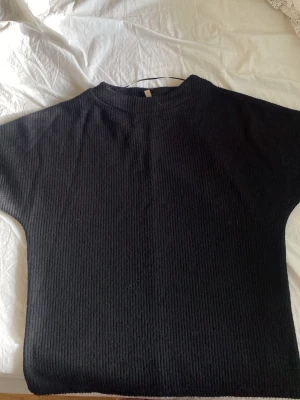 Svart ribbstickad tröja Soyaconcept - Svart ribbstickad tröja från Soyaconcept med rund halsringning och korta ärmar. Tröjan har en enkel och stilren design, perfekt för lager på lager och passar till många olika outfits. Materialet är mjukt och bekvämt.
