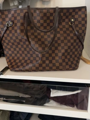 Brun Louis Vuitton Damier handväska - Säljer en klassisk brun handväska från Louis Vuitton i Damier-mönster. Väskan har två handtag, guldfärgade metalldetaljer och är tillverkad i canvas med läderdetaljer. Perfekt för dig som gillar tidlös och lyxig design. Eftersom jag köpt den secondhand så kan jag ej säga med säkerhet att den är äkta! Vet ej storleken på den heller!
