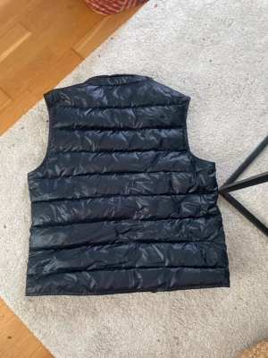 Mörkblå moncler dunväst från Moncler - Snygg mötkblå. dunväst från Moncler med klassisk quiltad design och glansigt material. Västen har Monclers ikoniska logotyp på bröstet och breda horisontella kanaler. Perfekt för lager-på-lager och streetstyle.