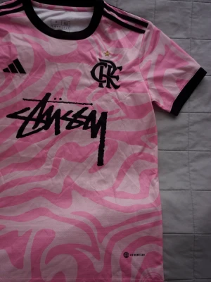 Rosa Stüssy x Adidas | Fotbollströja - Unik rosa sporttröja från Adidas i samarbete med Stüssy, med svart grafiskt mönster och logga på bröstet. Tröjan har svarta detaljer vid krage, ärmslut och axlar samt CR Flamengo-emblem. Tillverkad i lätt och ventilerande material. Tröjan är helt ny och väldigt limited! 
