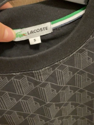 Mönstrad svart t-shirt från Lacoste - Snygg svart t-shirt från Lacoste med geometriskt mönster i grått. T-shirten har rund halsringning och gröna samt vita detaljer vid krage och ärmslut. Klassisk Lacoste-logga på bröstet. Perfekt för dig som gillar stilrena men unika plagg.