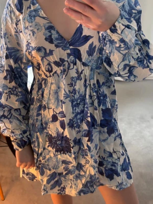 Blåvit blommig kortklänning - Supersöt kortklänning med blått blommönster på vit botten. Klänningen har v-ringning, långa puffärmar och en luftig passform. Perfekt för sommarens alla tillfällen och ger en fräsch och trendig look. Strl xs men passar mig som bär s-m