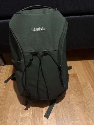 Mörkgrön ryggsäck från Haglöfs - Snygg mörkgrön ryggsäck från Haglöfs med minimalistisk design. Ryggsäcken har vadderade axelband, elastisk snörning på framsidan, sidoficka och diskret logga. Tillverkad i slitstarkt tyg, perfekt för skola eller utflykter.