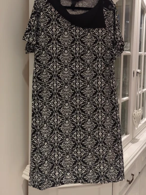 Svartvit mönstrad klänning från Jean - Kort svartvit klänning från Jean med elegant mönster och rund halsringning. Klänningen har korta ärmar och ett klassiskt, stilrent tryck som ger en snygg kontrast. Perfekt för dig som gillar grafiska detaljer och enkel design.