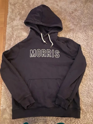 Svart hoodie från Morris - Snygg svart hoodie från Morris med vit logga broderad på bröstet. Klassisk känguruficka framtill, vita dragsnören i huvan och ribbade muddar vid ärmar och nederkant. Perfekt för en avslappnad och trendig stil.