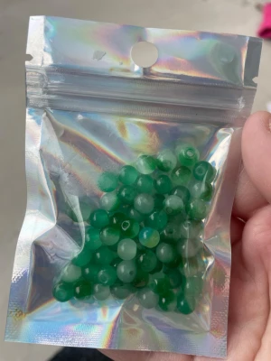 Gröna/vita glas pärlor i plastpåse - Säljer ett gäng runda gröna pärlor i en genomskinlig och holografisk plastpåse. Perfekta för att göra egna smycken som armband eller halsband. Pärlorna har en slät yta och hål i mitten för enkel trädning.