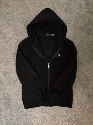 Svart hoodie från Polo Ralph Lauren - Snygg svart hoodie från Polo Ralph Lauren med dragkedja framtill och klassisk huva. Tröjan har det ikoniska vita Polo-märket broderat på bröstet och är tillverkad i mjukt bomullsmaterial. Perfekt för en avslappnad och stilren look. Skulle rekommendera tröjan till nån som har storlek S eller XS, tröjan är lite mindre en i storlek!