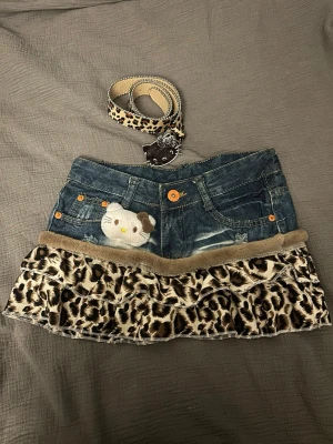 Jeanskjol med leopardvolanger och Hello Kitty - Unik jeanskjol med volanger i leopardmönster, matchande leopardbälte och Hello Kitty-detalj framtill. Kjolen har fickor, dekorativa sömmar och en söt Hello Kitty-nalle fastsydd på ena sidan. Perfekt för dig som gillar statement-plagg och djurmönster.
