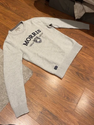 Ljusgrå stickad tröja från Morris - Snygg ljusgrå stickad tröja från Morris med rund halsringning och ribbade muddar. Framtill finns ett stort Morris-tryck och ett broderat emblem i blått och vitt. Perfekt för dig som gillar klassisk och stilren design.