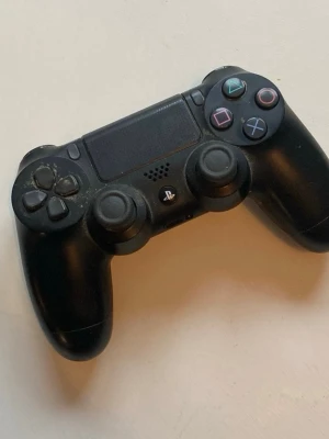 PlayStation 4 med DualShock 4 kontroll - Säljer en svart PlayStation 4 konsol från Sony tillsammans med en svart DualShock 4 kontroll. Konsolen har klassisk rektangulär form och flera anslutningar på baksidan, medan kontrollen har touchpad och färgade knappar. Perfekt för gaming och multiplayer. vi kan sänka priset