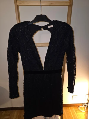 Svart glittrig klänning - Säljer en svart glittrig kortklänning med lång ärm och djup öppen rygg. Klänningen har en markerad midja och är täckt av glittriga detaljer som ger extra wow-faktor. Perfekt för dig som vill sticka ut och glänsa.